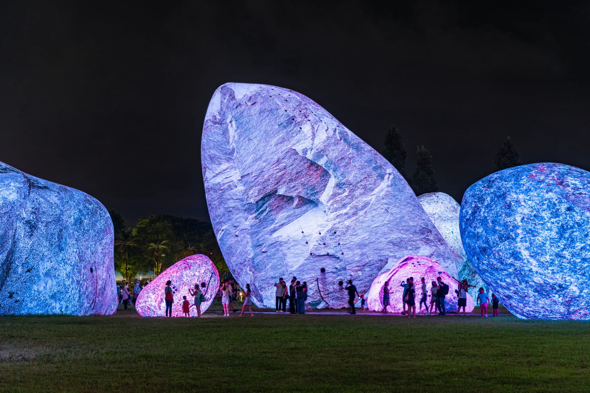 towering rocks rest in a park in glowing hues of purple and blue. passerby observe.