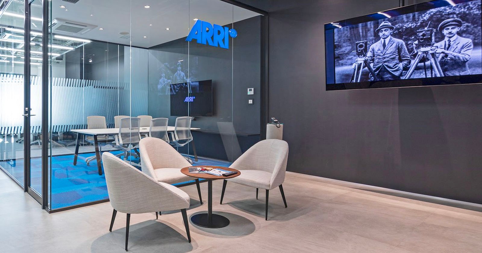 A modern office waiting area with three beige chairs and a small round table with magazines. A large glass-walled conference room is in the background. A flat-screen TV on the wall displays a black-and-white image of two people using early film equipment. The ARRI logo is visible.