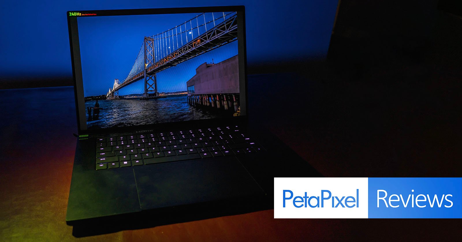 A laptop with an illuminated keyboard is open on a dark surface. The screen displays a vivid image of a bridge over water during twilight. "PetaPixel Reviews" is written in a blue and white banner at the bottom right corner. .