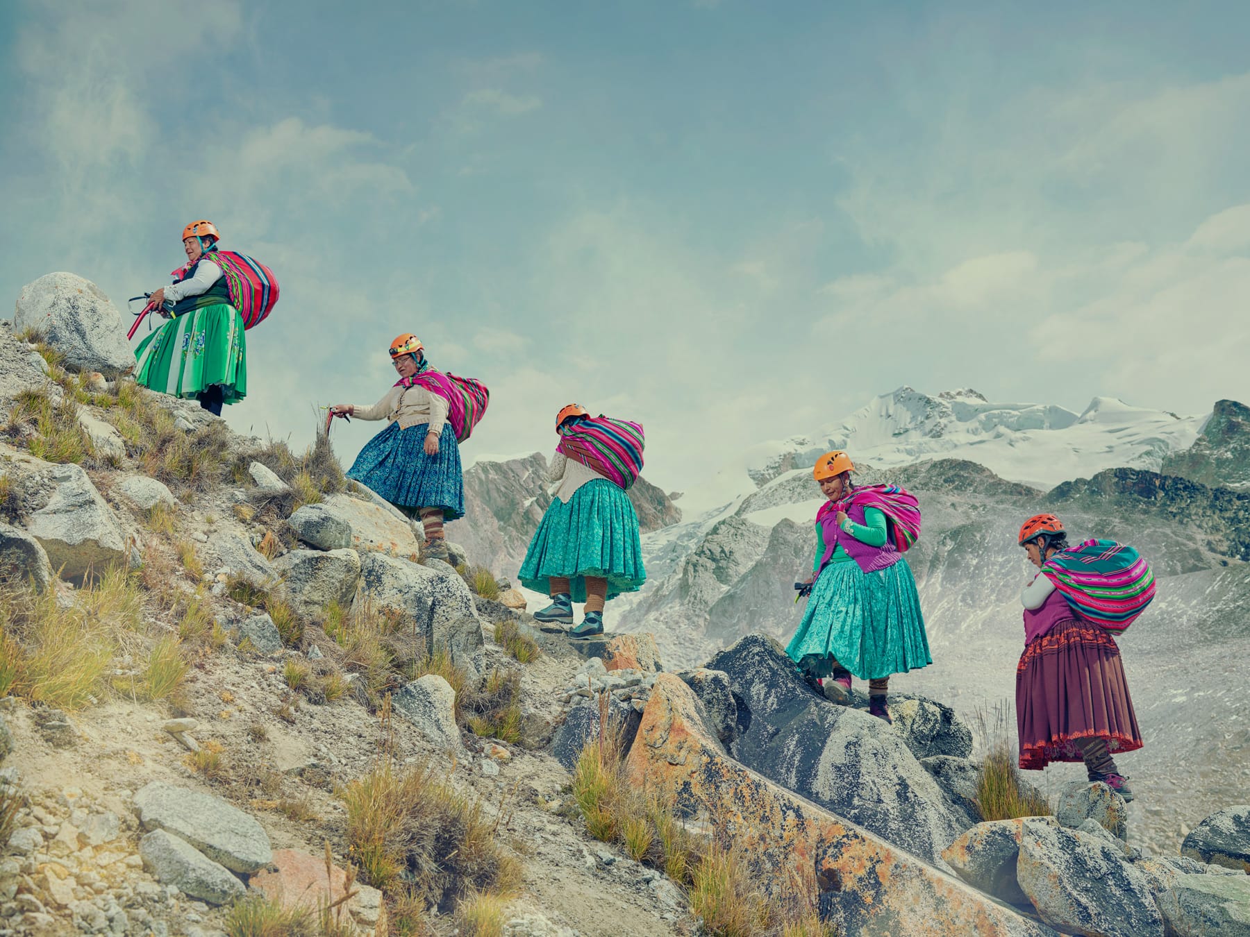 five Indigenous Bolivian "cholitas" climb the side of a mountain, wearing traditional billowing skirts, orange helmets, and using their shawls as rucksacks