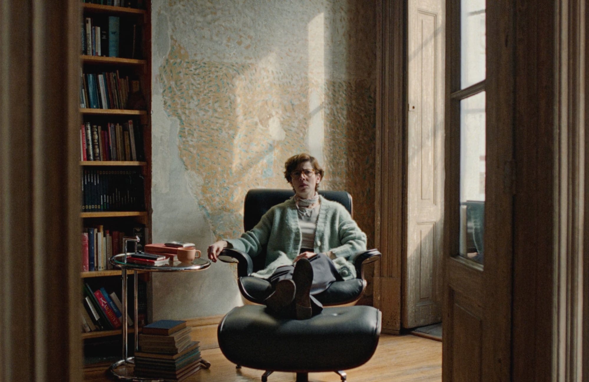 a white woman in glasses sits next to a small table and bookshelf with her feet up