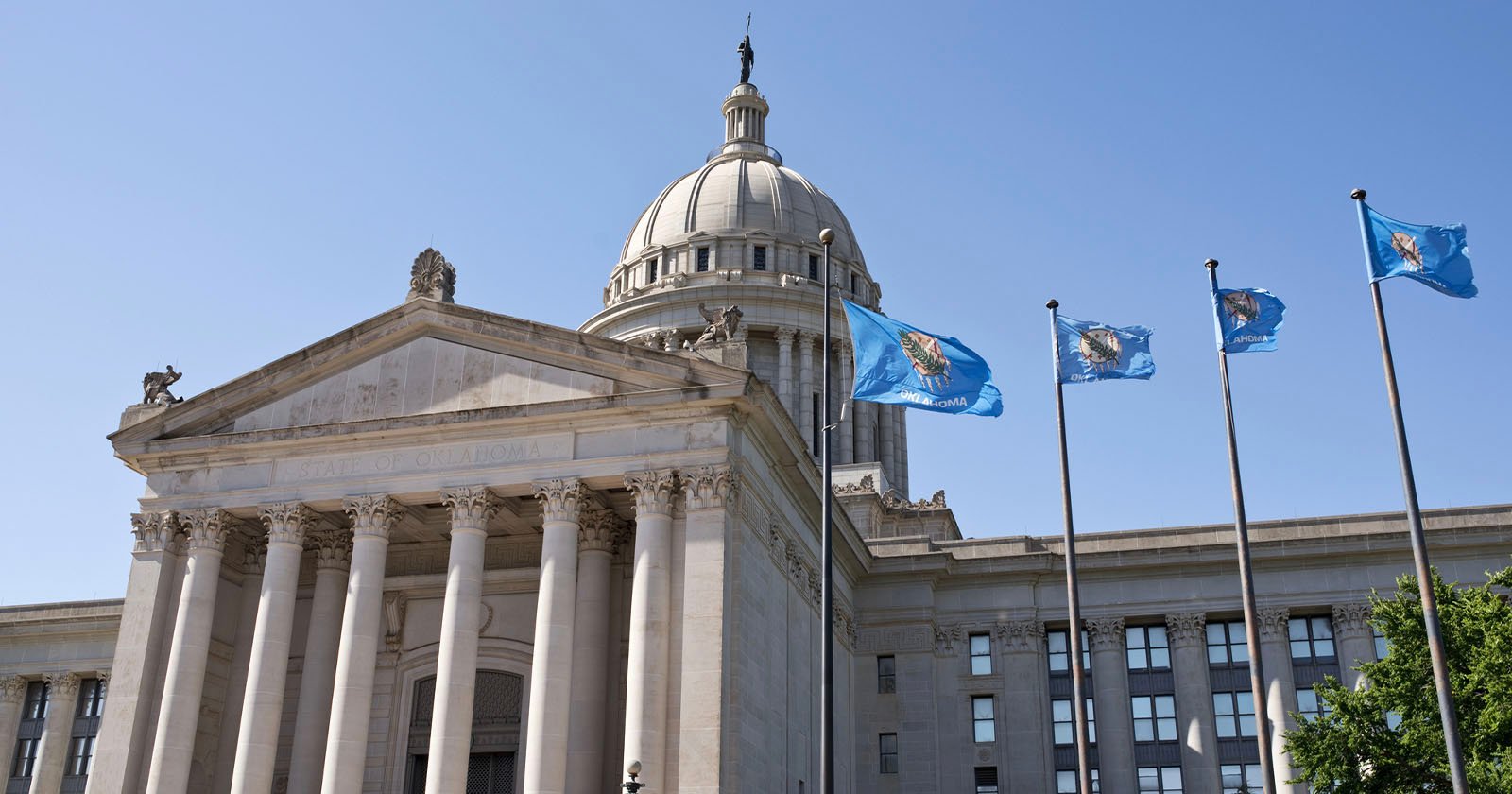 The image shows a grand neoclassical building with a large dome and columns, which is the Oklahoma State Capitol. Three blue flags with the state emblem are flying in the foreground against a clear blue sky. Green trees are partially visible on the side.