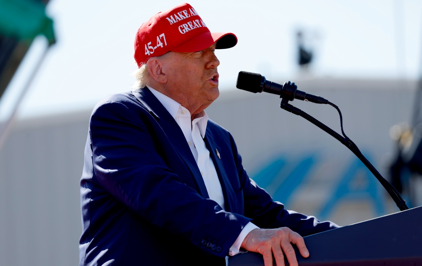 Former President Donald Trump at a podium on an outdoor stage, wearing a Make American Great Again hat.